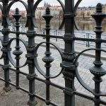 Forged metal railings on the Vltava river embankment in Prague. In the background is a view of Prague Castle with St. Vitus Cathedral on a hill above the river.