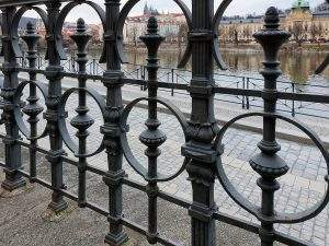 Forged metal railings on the Vltava river embankment in Prague. In the background is a view of Prague Castle with St. Vitus Cathedral on a hill above the river.