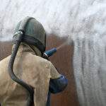 A man weating full safety gear uses ground glass to sandblast the steel casing of a meat drying machine for a rendering plant